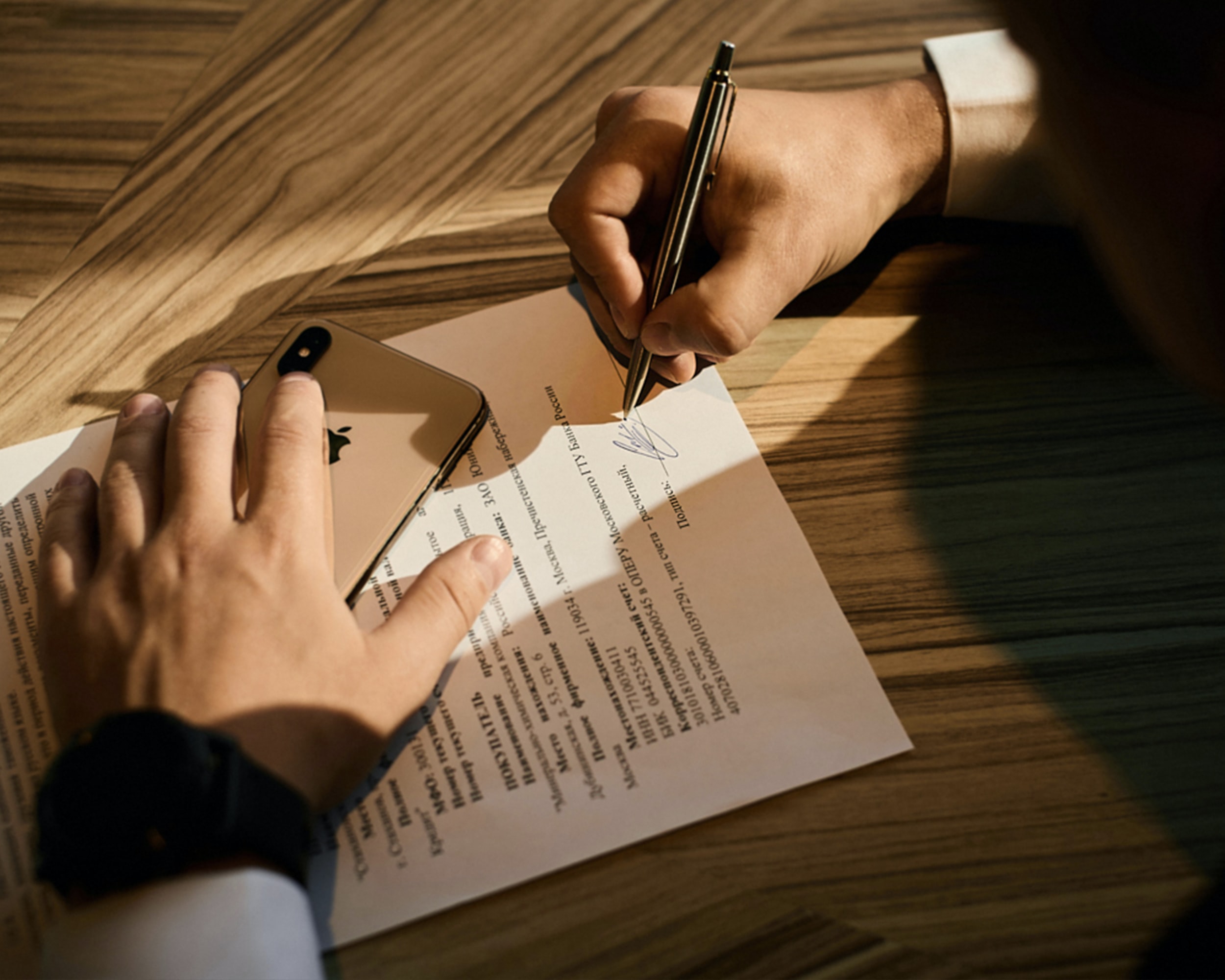 Person holding gold mobile phone in one hand while signing a document with the other