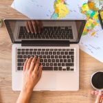 A birds eye view of a laptop surrounded by maps on a table being used by a person holding coffee