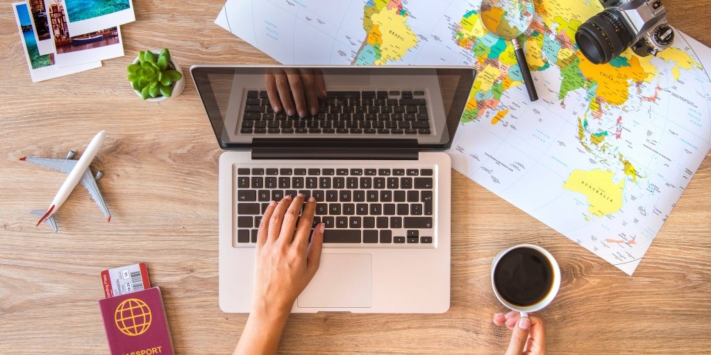 A birds eye view of a laptop surrounded by maps on a table being used by a person holding coffee